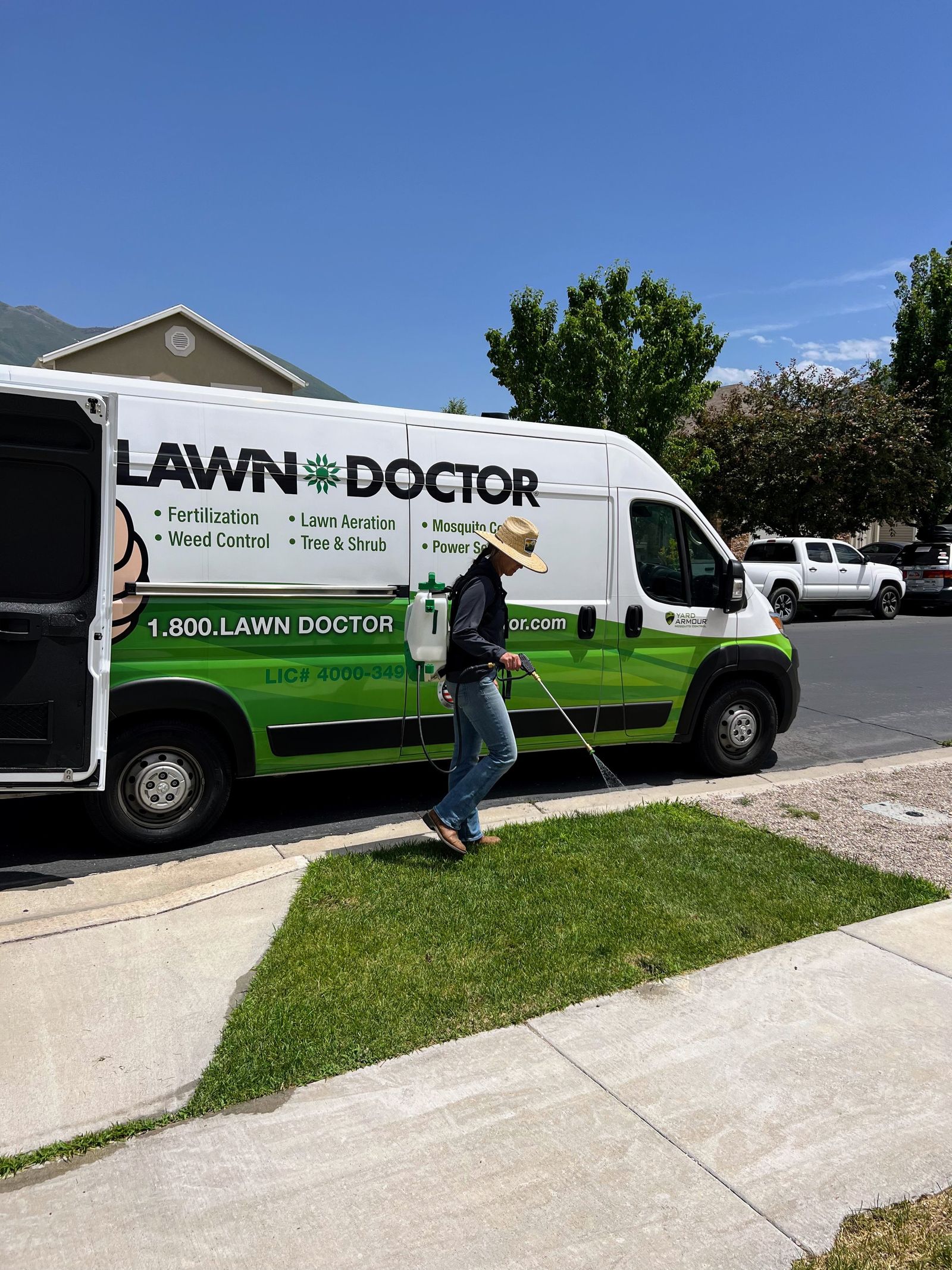 Lawn Doctor technician in a straw hat spraying a treatment next to a green and white service vehicle.